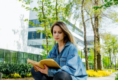 Young student girl in denim jacket reading a book at alley near modern building of University.