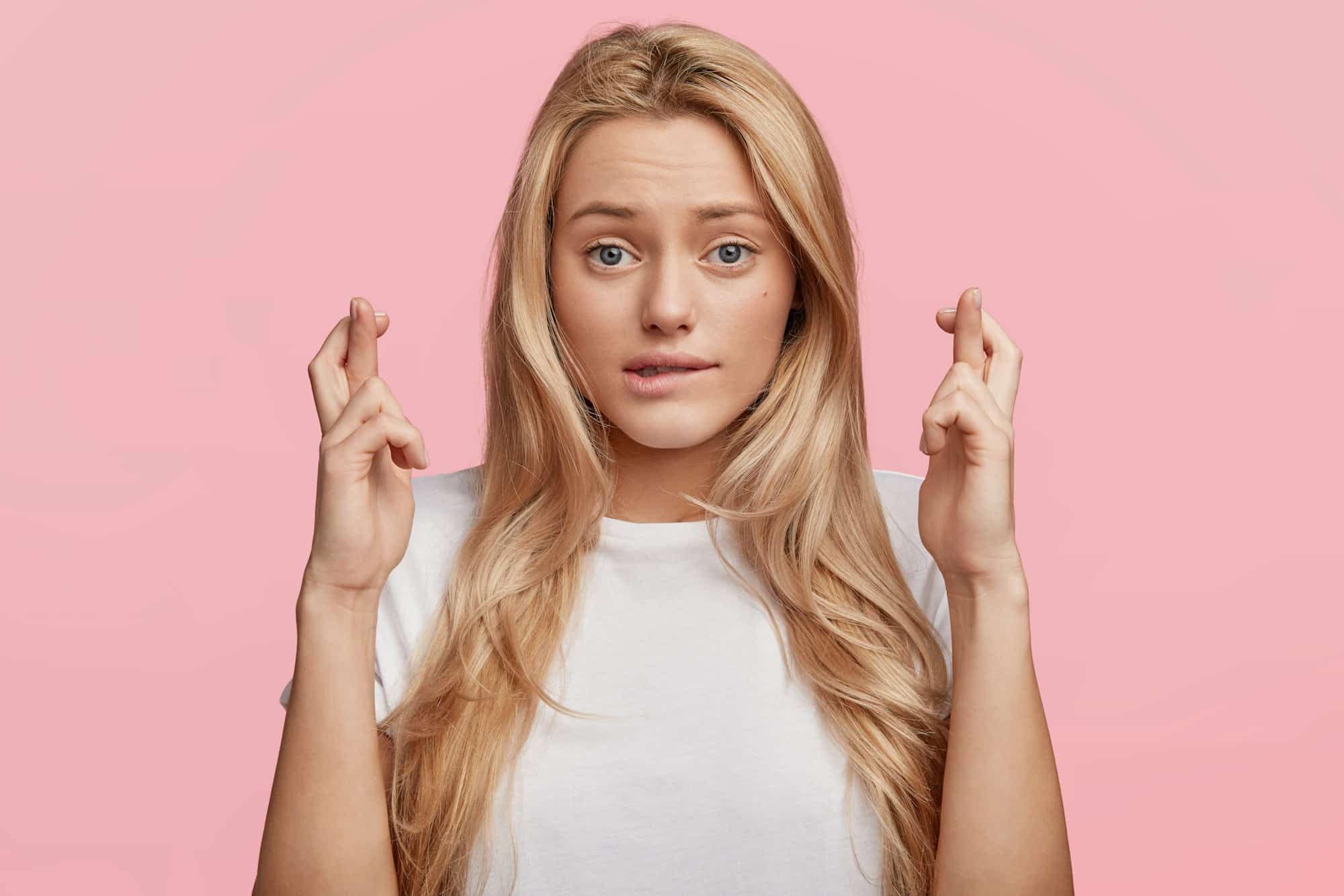 Studio shot of blonde lovely female student has hopeful expression, crosses fingers and bites lower