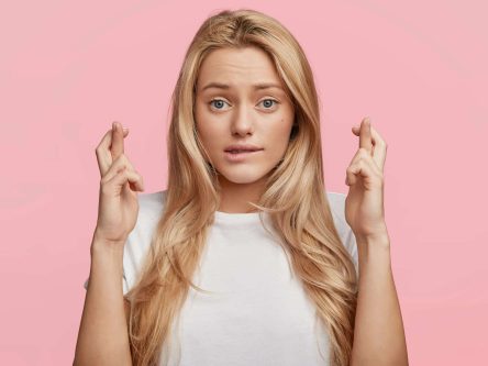 Studio shot of blonde lovely female student has hopeful expression, crosses fingers and bites lower